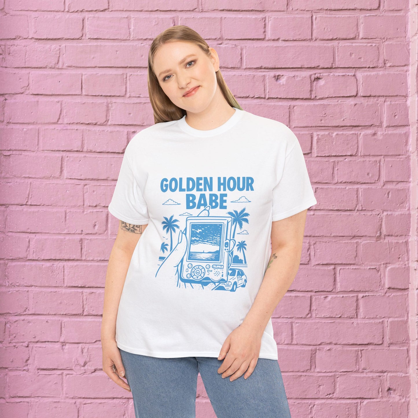 Person wearing a white t-shirt with 'Golden Hour Babe' design against a pink brick wall.