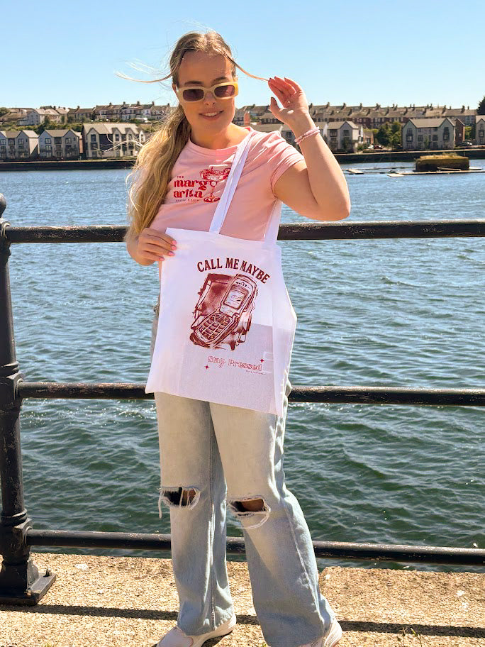 Person holding a 'Call Me Maybe' tote bag by the water