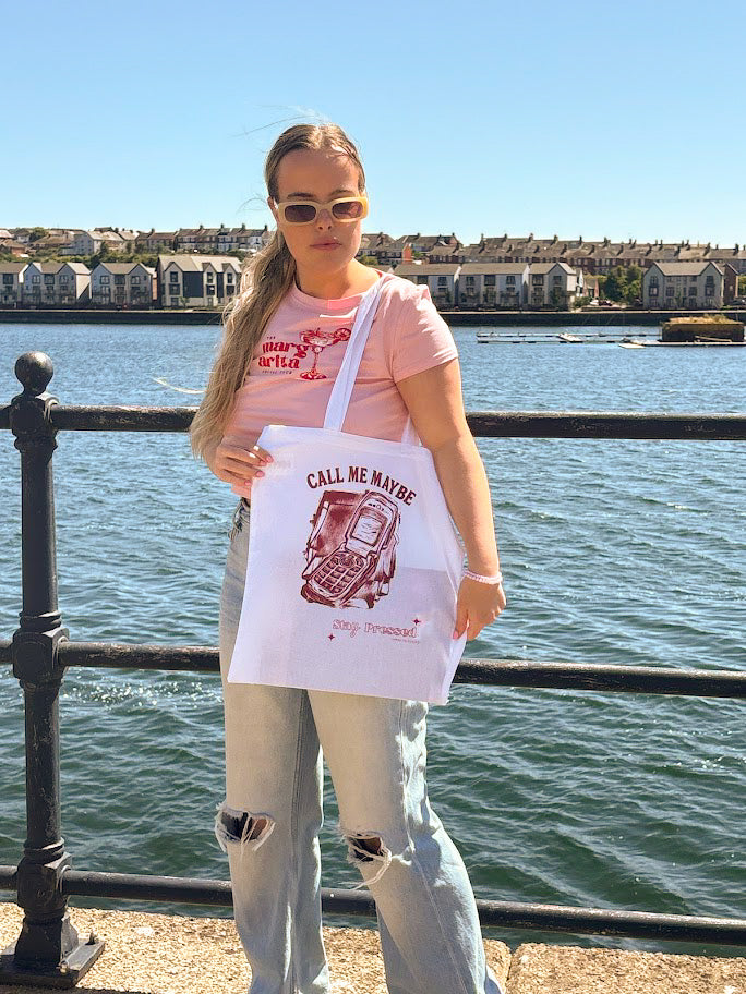 Person holding a 'Call Me Maybe' tote bag by a waterfront with houses in the background
