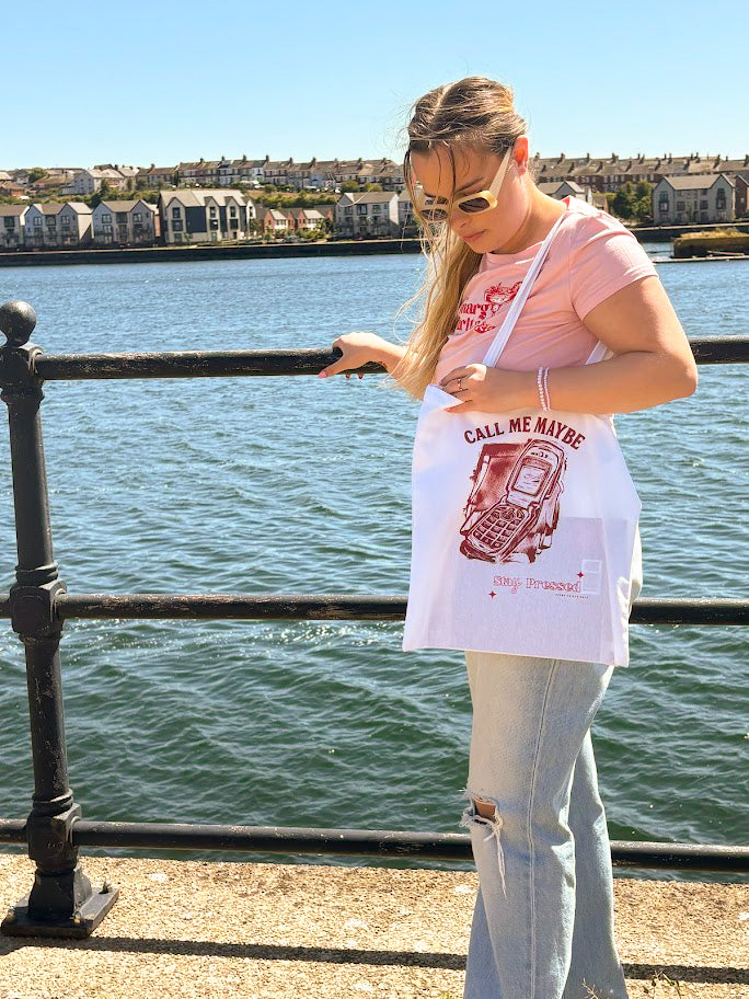 Woman holding a 'Call Me Maybe' tote bag by the water