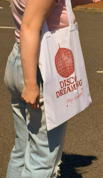 Person holding a tote bag with 'Disco Dreaming' text and red graphic.