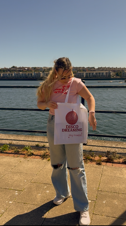 Person holding a tote bag with 'Disco Dreaming' text by a waterfront.